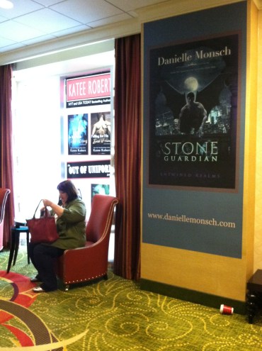 Lobby area wall and window posters, floor to ceiling, with fantasy and suspense book covers, looming over conference attendee seated in armchair.
