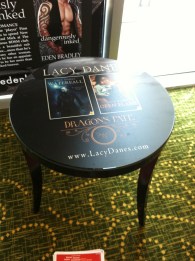 Occasional table in lobby area, with laminated image of Lacy Danes book covers; images of fantasy heroes with tattoos and leather jackets